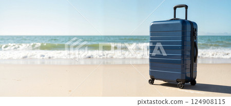 Blue suitcase stands on sandy beach, ocean waves in background. Sunlight reflects off metallic surface. Seashore suggests travel or vacation mood, invoking adventure 124908115