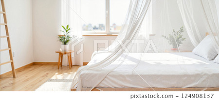 Minimalist bedroom with sheer canopy over white bed, light wooden floor, ladder, small table with plant, and large window. Soft, natural light fills the room, creating an airy feel 124908137