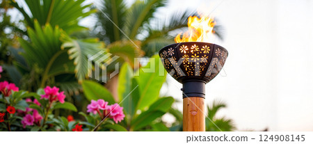 Decorative metal torch with floral cutouts and flames complements vibrant pink flowers and lush greenery, creating a tropical garden ambiance, evoking warmth and elegance 124908145