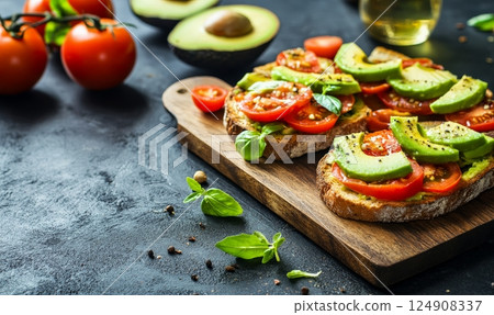 Slices of bread topped with avocado and tomato on a wooden board, surrounded by fresh ingredients. 124908337