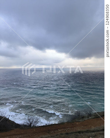 Dramatic sky with large grey clouds above choppy ocean waves, viewed from a grassy cliff edge. The sea has a gradient of colors from deep blue to lighter teal near the shore 124908350