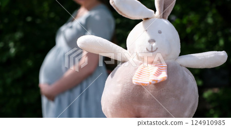 Soft plush bunny toy in focus with a blurred pregnant woman holding her belly in the background Soft plush bunny toy in focus with a blurred pregnant woman holding her belly in the background 124910985