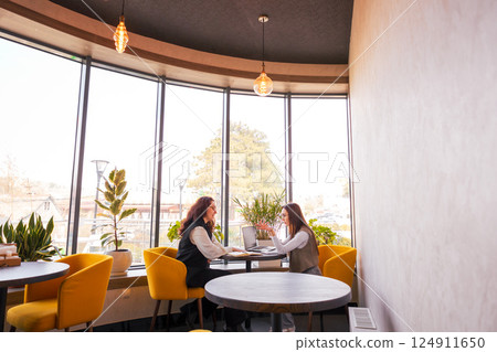 Two smartly dressed young Caucasian women sitting at a cafe and having a deep conversation about marketing strategies. Concept of online business growth and professional cooperation. Wide shot 124911650