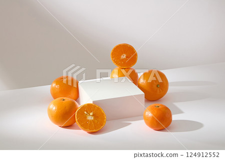 Half an orange placed upright on another ripe orange. White square pedestal with empty space for product placement. Oranges displayed around the podium, presented on a minimalist white background.  124912552