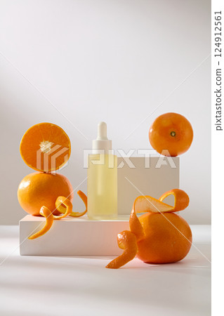 Composition of a dropper bottle placed on a rectangular white podium. Orange placed at the edge of the square podium. Half of orange is placed on others, next to peel cut into wavy strips. 124912561