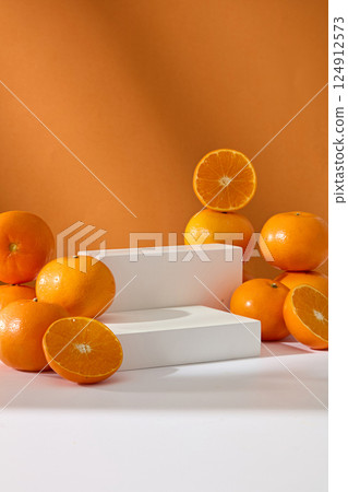 Vertical photo of two white podiums placed side by side like steps, to display products. Ripe oranges with shiny, vibrant peel. Oranges cut in half stacked on top of each other and placed around. 124912573