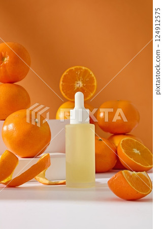 Glass dropper bottle containing essence extracted from fresh oranges. Tropical oranges are placed on top of each other and displayed on white podium. Orange peel is sliced to thin waves. 124912575