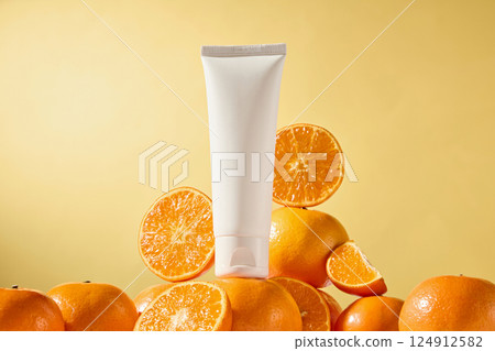 Unlabeled tube of cream stands on oranges, fresh oranges cut in half in behind, showing their juicy flesh. Light shines in citrus with shiny soft peel. Light brown background. Advertising photo. 124912582