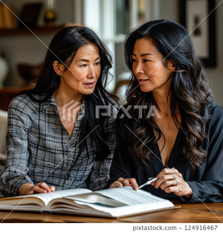 Two women are sitting at a table looking at a book 124916467