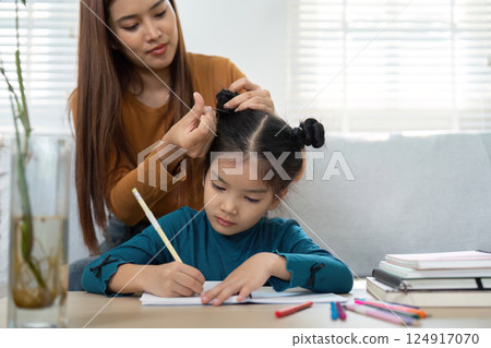 Bonding Through Education and Grooming. A parent helps a child with hair while she focuses on her homework. 124917070