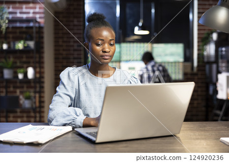 Determined female entrepreneur analyzing financial data on laptop at her desk, surrounded by trading documents. Her coworker, slightly blurred, works in the background at a multiscreen setup. Determined female entrepreneur analyzing financial data on laptop at her desk, surrounded by trading documents. Her coworker, slightly blurred, works in the background at a multiscreen setup. 124920236