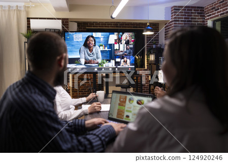 Multiethnic colleagues analyzing reports in late night meeting. Digital screen shows a videoconference call in progress, highlighting remote collaboration and business strategy in modern workspace. Multiethnic colleagues analyzing reports in late night meeting. Digital screen shows a videoconference call in progress, highlighting remote collaboration and business strategy in modern workspace. 124920246