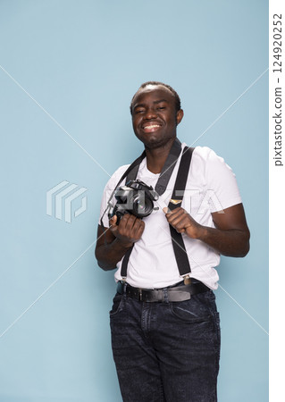 Young man in a photography studio wearing suspenders and a white t shirt. African american male individual smiles while holding his camera, exuding creativity and professionalism in a vibrant setting. Young man in a photography studio wearing suspenders and a white t shirt. African american male individual smiles while holding his camera, exuding creativity and professionalism in a vibrant setting. 124920252