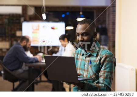 Professional black man deeply engaged in project planning, reviews financial analytics on his laptop. Behind him, coworkers discuss business trends, creating a dynamic workplace scene. 124920293
