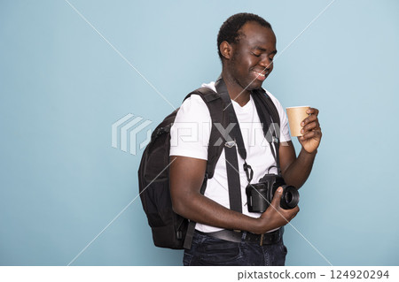 African American man touches his camera lens, smiling as he looks at the disposable cup in his hand. Black male photographer with a backpack and DSLR gazes at his coffee with a content expression. African American man touches his camera lens, smiling as he looks at the disposable cup in his hand. Black male photographer with a backpack and DSLR gazes at his coffee with a content expression. 124920294