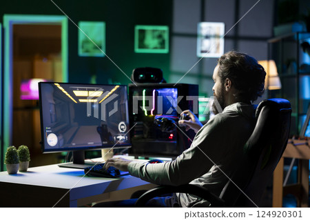 Man in dark living room playing videogame on gaming PC at computer desk, relaxing by shooting enemies, putting on headphones. Gamer wearing headset battling robots in singleplayer shooter 124920301