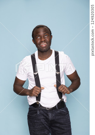 African American male model wearing suspenders, adjusting the straps while looking up with a smile. Black man grips the elastic suspenders holding his trousers as he stands against isolated backdrop. African American male model wearing suspenders, adjusting the straps while looking up with a smile. Black man grips the elastic suspenders holding his trousers as he stands against isolated backdrop. 124920305