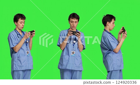 Medical assistant playing mobile video games on smartphone app, taking a break from healthcare work. Nurse enjoys gaming tournament on phone, standing against greenscreen backdrop. Camera B. Medical assistant playing mobile video games on smartphone app, taking a break from healthcare work. Nurse enjoys gaming tournament on phone, standing against greenscreen backdrop. Camera B. 124920336