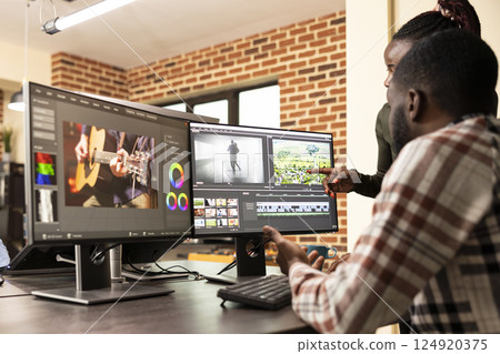 Professional male editor refining frames as coworker provides visual improvement support. Black man improving film quality at multi monitor workstation with assistance from female creative manager. 124920375