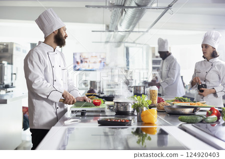 Diverse cooks cutting greenery and fresh vegetables with a knife, preparing ingredients for a new gourmet dish in restaurant kitchen. Professional culinary techniques for a fine dining experience. 124920403