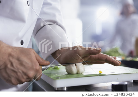 Sous chef pressing knife on cloves to make crushed garlic on the cutting board, creating condiments with a fresh flavor. Young cook preparing a gourmet dish at the counter, chef life. Close up. Sous chef pressing knife on cloves to make crushed garlic on the cutting board, creating condiments with a fresh flavor. Young cook preparing a gourmet dish at the counter, chef life. Close up. 124920424