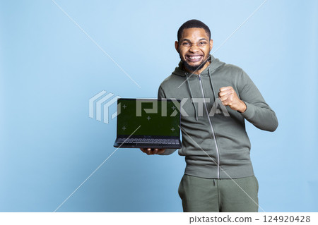 Confident happy person pointing at green screen mockup in studio, using personal laptop for an advertisement on camera. African american young guy gesturing against blue background. 124920428