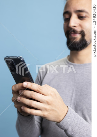 Confident thoughtful man working on his smartphone while engaging in digital communication, standing against blue background. Young casual adult doing remote work on modern online platforms. Confident thoughtful man working on his smartphone while engaging in digital communication, standing against blue background. Young casual adult doing remote work on modern online platforms. 124920430