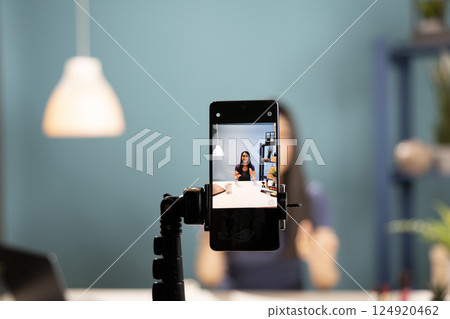 A smartphone camera captures a beauty influencer reviewing cosmetics. Mobile device screen showing asian vlogger using expressive gestures as she explains beauty products in a blue background studio. 124920462