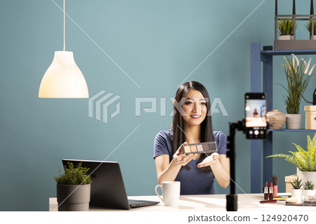 Cheerful asian woman holds an eyeshadow palette up to her phone camera, livestreaming a cosmetics review session. She shows makeup techniques, demonstrating products in an online makeup tutorial. Cheerful asian woman holds an eyeshadow palette up to her phone camera, livestreaming a cosmetics review session. She shows makeup techniques, demonstrating products in an online makeup tutorial. 124920470