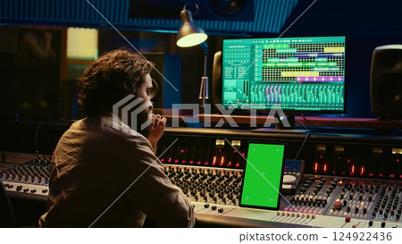 Music producer recording, mixing and reproducing sound on audio software, using stereo console and switchers to increase volume level and enhance tunes. Greenscreen display on tablet. Camera B. Music producer recording, mixing and reproducing sound on audio software, using stereo console and switchers to increase volume level and enhance tunes. Greenscreen display on tablet. Camera B. 124922436