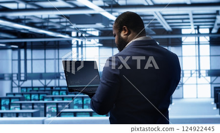 Computer scientist in server farm using laptop to patch network issues leading to slow data transfer rates. IT expert in data center establishment fixing errors affecting supercomputers, camera A 124922445