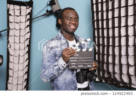 African American male director posing confidently in a studio with professional lighting, holding a clapperboard and DSLR. Black photographer smiling at camera with a film slate in his hands. 124922463
