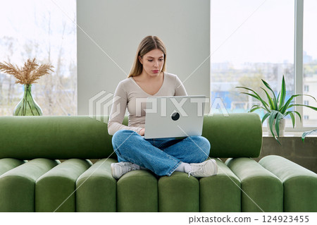 Portrait of young woman sitting on sofa with laptop, in home interior 124923455