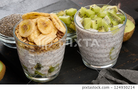 Two glasses of chia seed pudding, one topped with diced kiwi and the other with granola and banana chips, are on a dark surface with ingredients Two glasses of chia seed pudding, one topped with diced kiwi and the other with granola and banana chips, are on a dark surface with ingredients 124930214