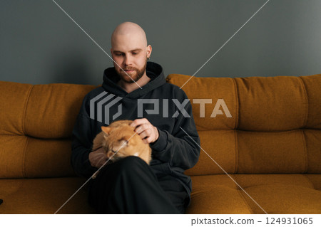 Portrait of young bald man with beard wearing casual clothes sitting on comfortable sofa, lovingly petting ginger cat, enjoying quiet moment at home. Concept of relationship between people and animal. Portrait of young bald man with beard wearing casual clothes sitting on comfortable sofa, lovingly petting ginger cat, enjoying quiet moment at home. Concept of relationship between people and animal. 124931065