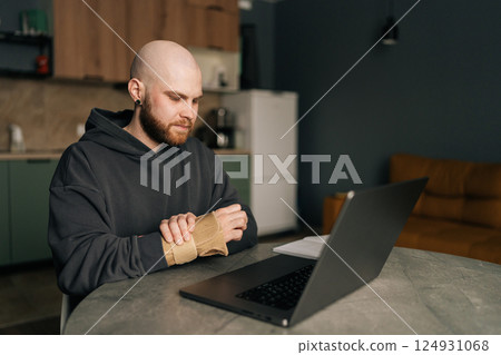 Sick freelancer programmer with wrist brace rubbing painful hand, working on laptop during carpal tunnel syndrome management at home workspace, sitting at table on background of kitchen interior. 124931068