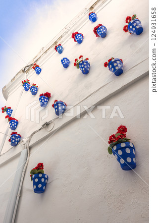 Cordoba town in Spain. Vibrant red flowers in blue polka-dot pots adorn a whitewashed wall, creating a cheerful and picturesque scene. 124931658