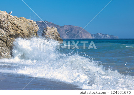 Nerja town, a resort of The Costa del Sol seaside region in Andalusia in the south of Spain. Ocean waves crashing against coastal rocks on a sunny day. A bird perched on a rock observes the scene. 124931670