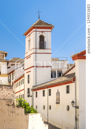 Granada town in Spain. Whitewashed building with red trim and bell tower against a vibrant blue sky. A charming architectural detail in a sunny, historical setting. Granada town in Spain. Whitewashed building with red trim and bell tower against a vibrant blue sky. A charming architectural detail in a sunny, historical setting. 124931683