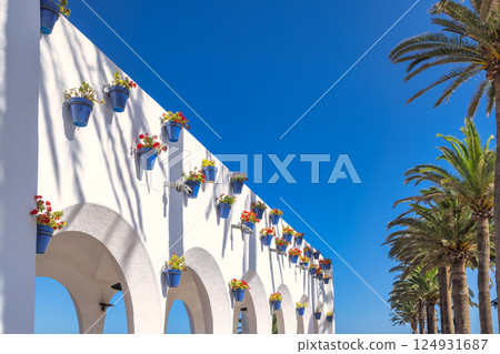 Nerja town, a resort of The Costa del Sol seaside region in Andalusia in the south of Spain. Vibrant flowers in blue pots adorn a white archway wall, set against a backdrop of palm trees. 124931687