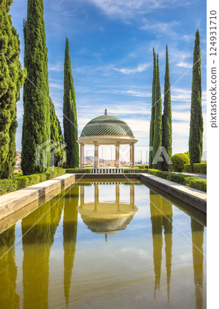 The Historical Botanical Garden La Concepcion in Malaga city at Andalusia, Spain, Europe. Serene gazebo reflected in a tranquil pond, surrounded by lush greenery and tall cypress trees. 124931710