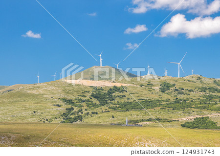 View of country in Bosnia and Herzegovina, Europe. Wind turbines generate clean power atop a rolling green landscape under a sunny blue sky with scattered clouds. Eco-friendly energy concept. 124931743