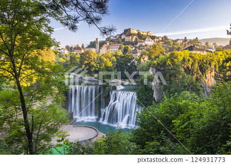 The Pliva Waterfall in Jajce town, Bosnia and Herzegovina, Europe. Cascading waterfall surrounded by lush greenery, with a historic town and fortress perched atop a hill in the background. 124931773
