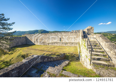 Walled City of Jajce, medieval fortress in Jajce town in Bosnia and Herzegovina, Europe. Stone fortress walls and stairway under clear blue sky, lush green hillside. 124931781