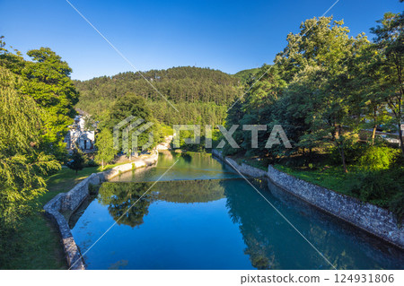 The Pliva river in Jajce town in Bosnia and Herzegovina, Europe. Scenic river view with stone embankments surrounded by lush green trees and a forested hillside under a clear blue sky. 124931806