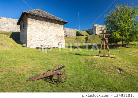 Walled City of Jajce, medieval fortress in Jajce town in Bosnia and Herzegovina, Europe. Scenic view of historical site featuring a stone building, castle wall, wooden cannon, and vibrant green lawn. 124931813