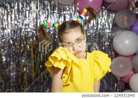Offended girl in glasses and yellow holiday blouse with funny ponytails hairstyle on festive background with balloons 124931840