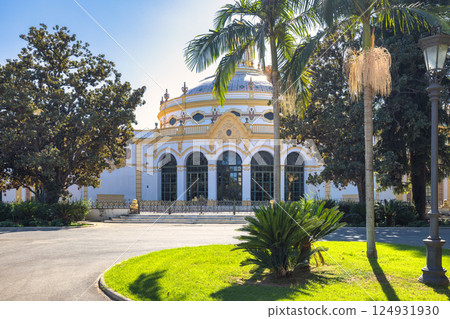 Seville town, capital of Andalusia in Spain. Elegant rotunda building in a sunny park setting, surrounded by lush greenery and palm trees. 124931930