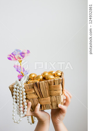 Child holding wicker basket with gold eggs, beads, and pink flower 124932861