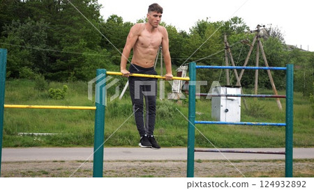 Hardy shirtless sportsman doing push ups on horizontal bar at nature. Strong and muscular guy training at sports ground. Young athletic guy exercising outdoor. Concept of active and healthy lifestyle Hardy shirtless sportsman doing push ups on horizontal bar at nature. Strong and muscular guy training at sports ground. Young athletic guy exercising outdoor. Concept of active and healthy lifestyle 124932892